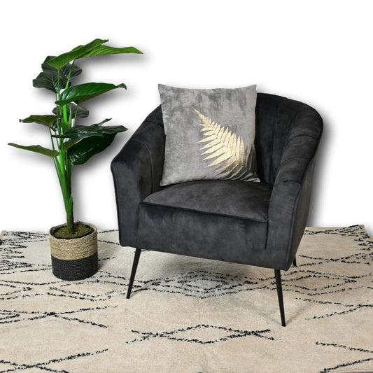 Donkergrijze fluwelen stoel met goud bladkussen, naast een groene plant in een mand op een beige tapijt.