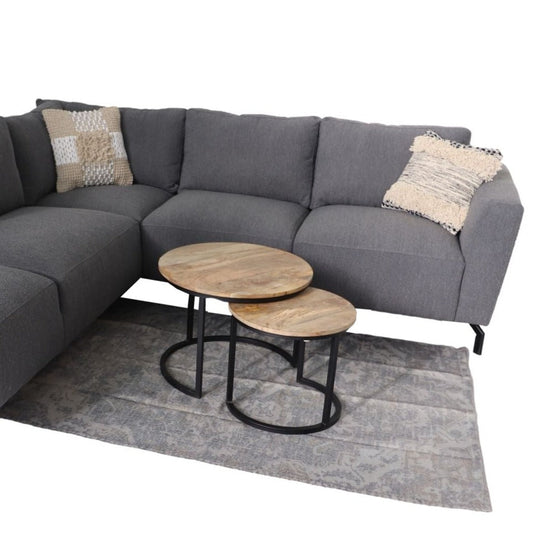 Donkergrijze hoekbank met twee sierkussens, naast houten salontafels en een subtiel patroon vloerkleed.