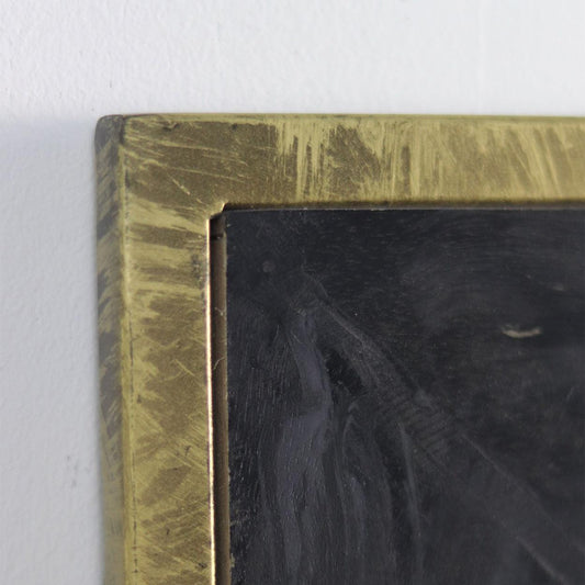 Detail van een zwart bord met robuuste, verweerde messing omlijsting tegen een lichte muur.