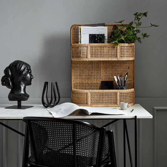 Zwart borstbeeld, open boek, en rieten organizer met groen op een witte tafel tegen een grijze muur.