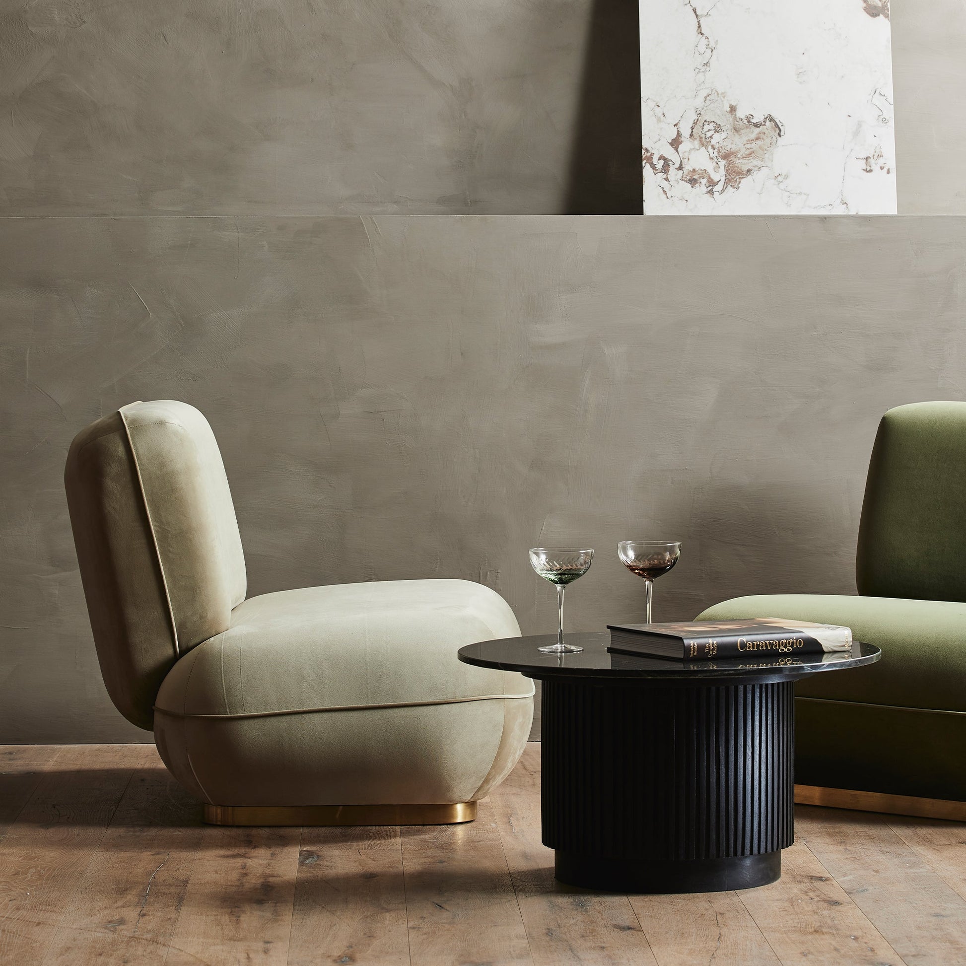 Twee fluwelen fauteuils in beige en groen naast een zwarte salontafel met glazen en een boek op houten vloer.