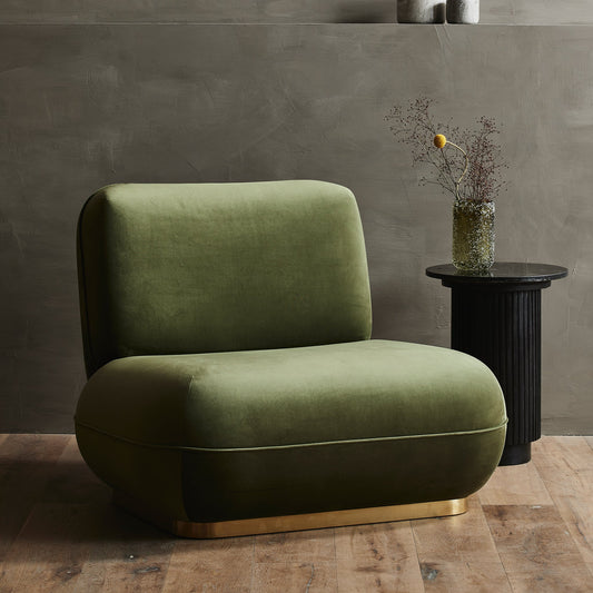 Groene fluwelen fauteuil met gouden basis naast zwart bijzettafeltje met vaas en takken, op houten vloer.