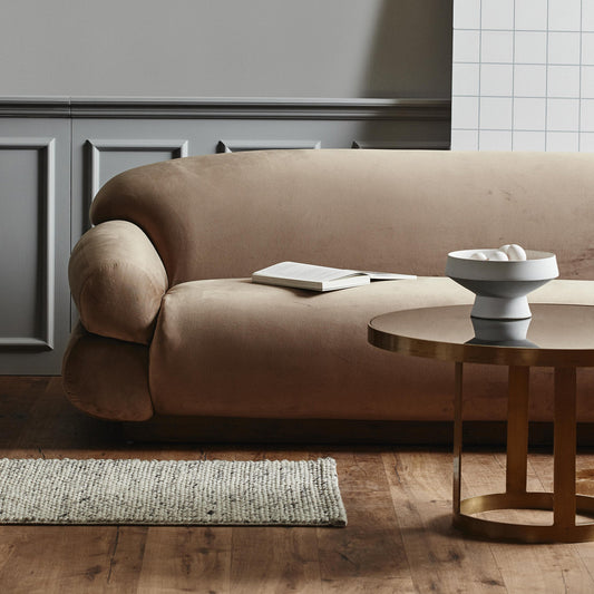 Zachte beige bank met open boek, naast een ronde houten tafel met witte schaal op houten vloer.