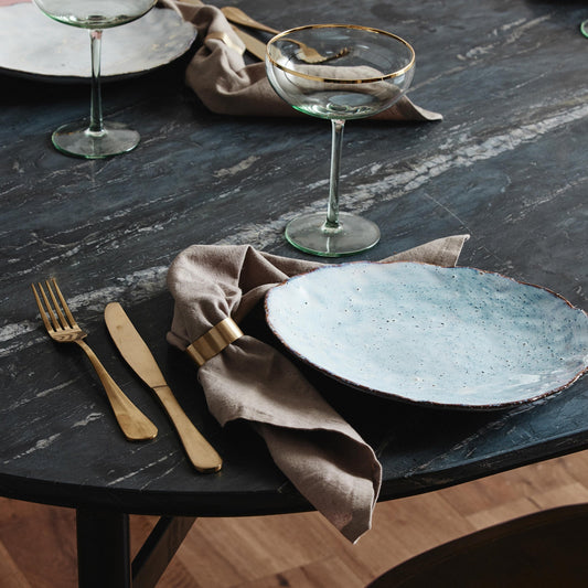 Elegante tafelschikking met blauw keramisch bord, gouden bestek en glazen coupe op donkere marmeren tafel.