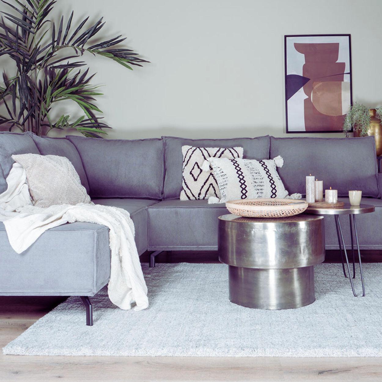 Grijze hoekbank met kussens, gestapelde ronde metalen tafels, kaarsen en abstracte muurkunst in een moderne woonkamer.