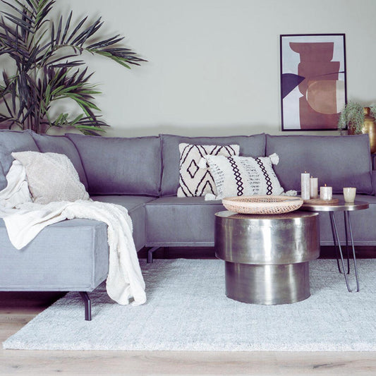 Grijze hoekbank met sierkussens, metalen salontafels en kaarsen, tegen een neutrale muur met abstract schilderij.