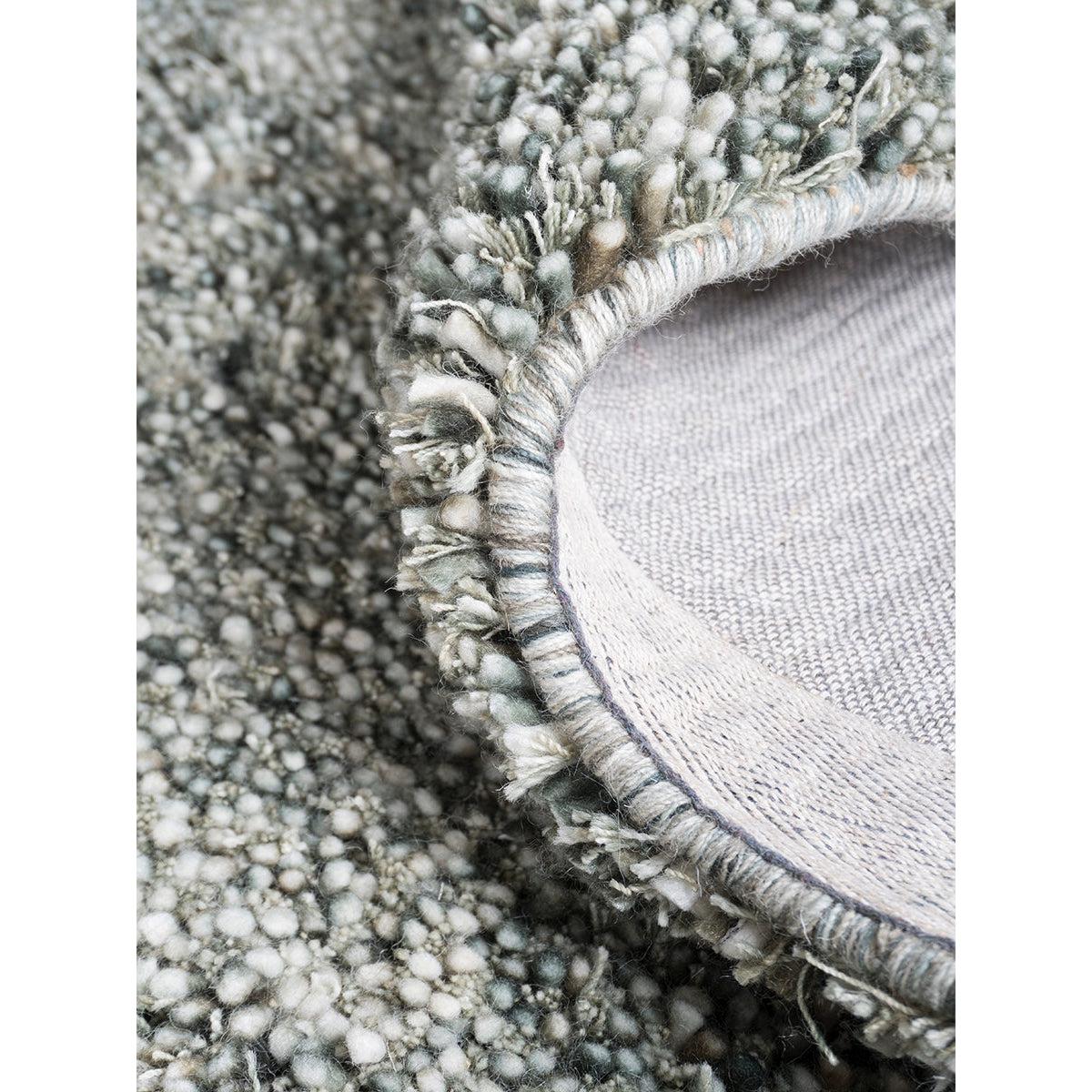 Close-up van een geweven wollen tapijt met gedetailleerde rand, in grijsgroene tinten en zachte textuur.