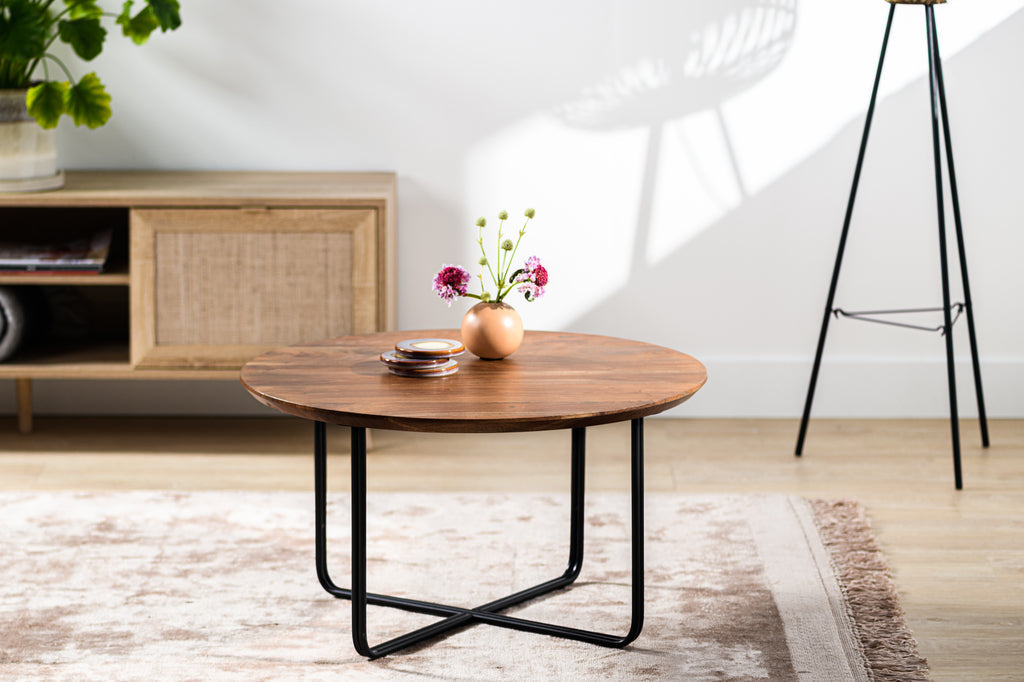 Ronde houten salontafel met zwarte poten, een vaas met bloemen en stapel schoteltjes, op lichtgekleurd tapijt.