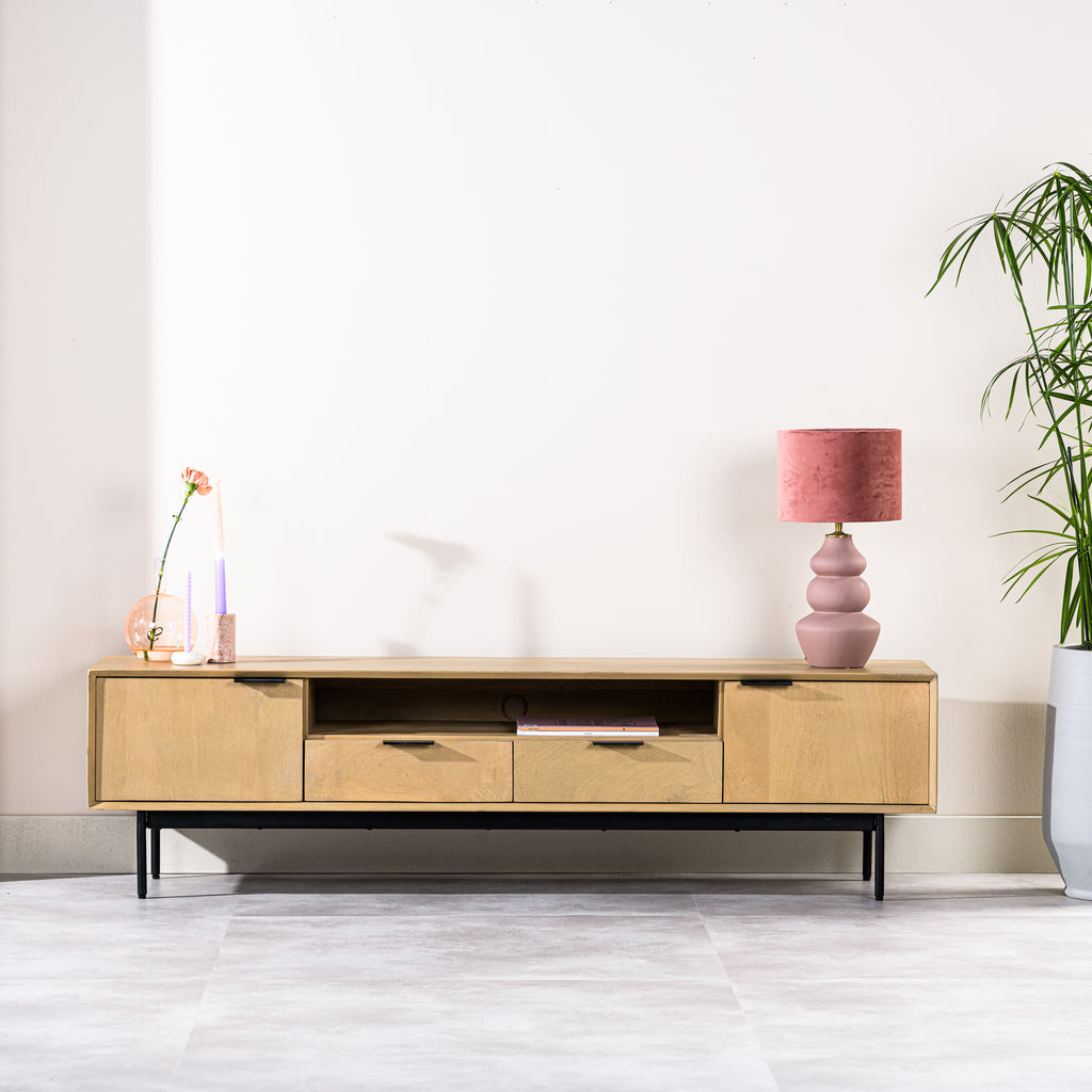 Licht houten dressoir met roze lamp, vaas met roze bloem en twee kaarsen, naast groene plant.