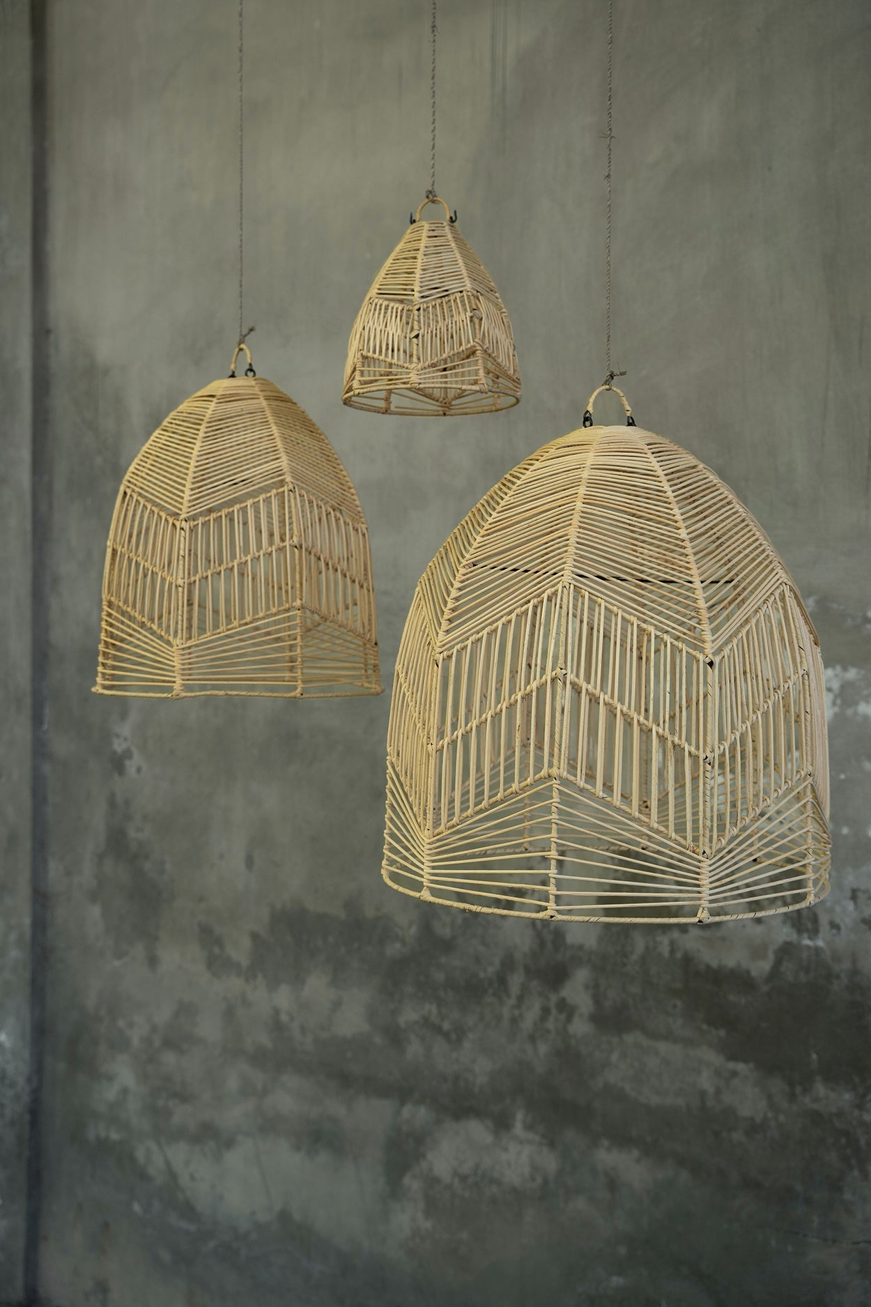 Drie hanglampen van gevlochten riet tegen een betonnen muur, variërend in grootte en vorm.
