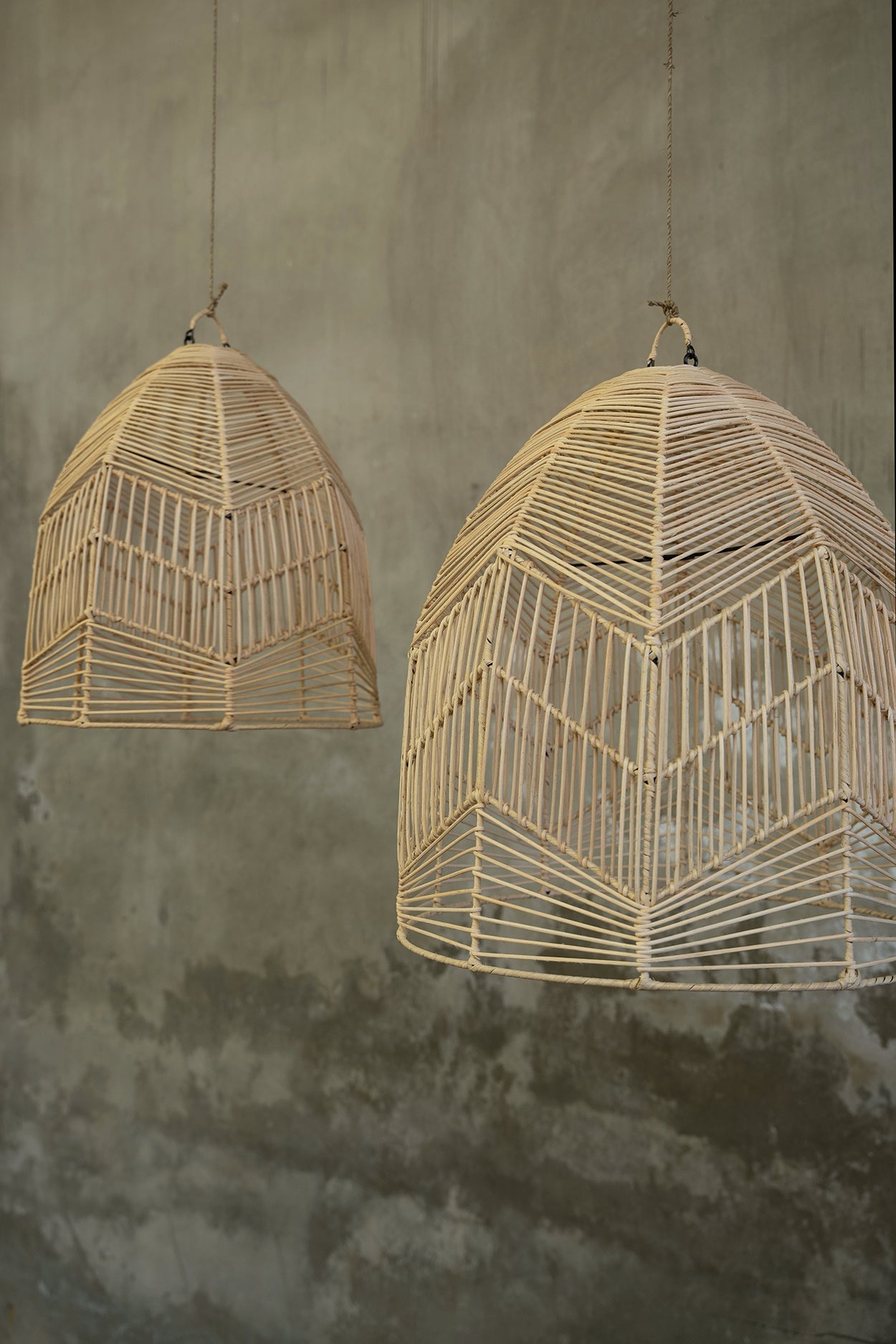Twee handgemaakte rotanlampen met geometrisch patroon hangen voor een grijze ruwe muur.