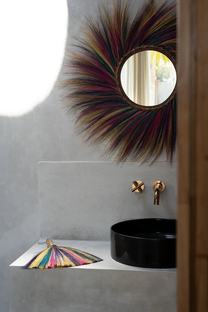 Kleine grijze badkamer met kleurrijke ronde spiegel en zwarte wasbak.  Gouden kraan met een decoratieve waaier.
