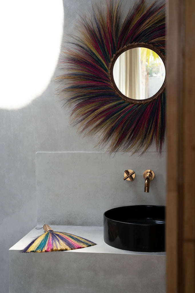 Ronde spiegel met regenboogkleurige rand boven zwarte wasbak en gouden kraan in een minimalistische badkamer.