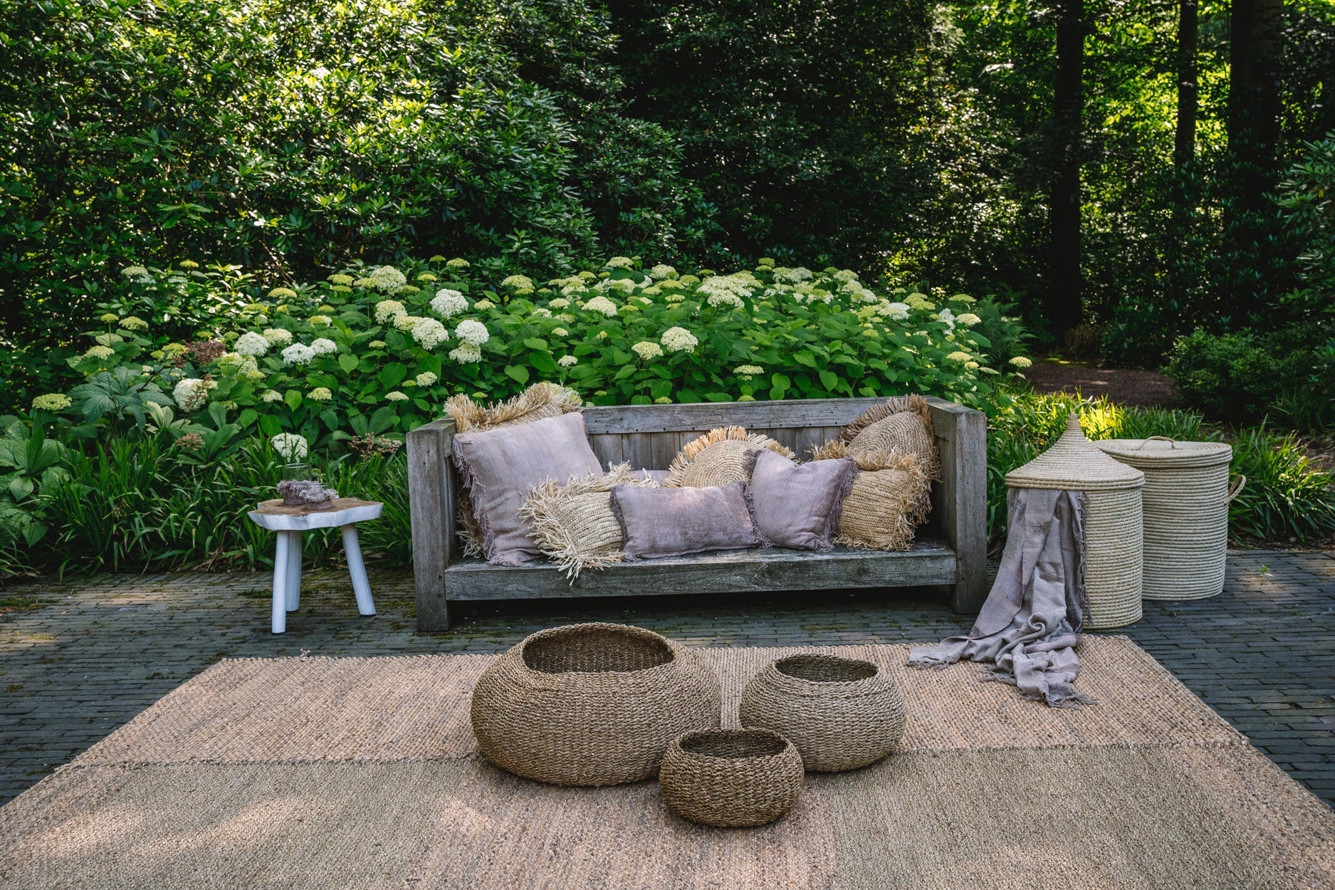 Houten bank met kussens op een patio, omgeven door groene planten en rieten manden. Rustieke buitenstijl.