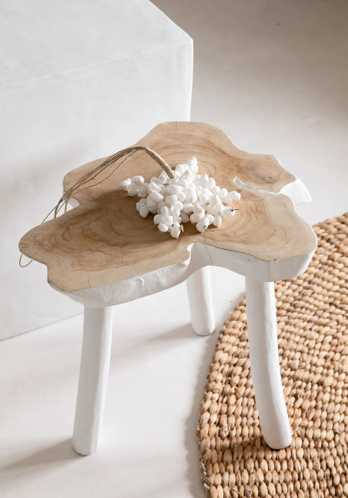 Kleine houten kruk met witte tros decoratieve schelpen, naast een rond gevlochten juten tapijt.