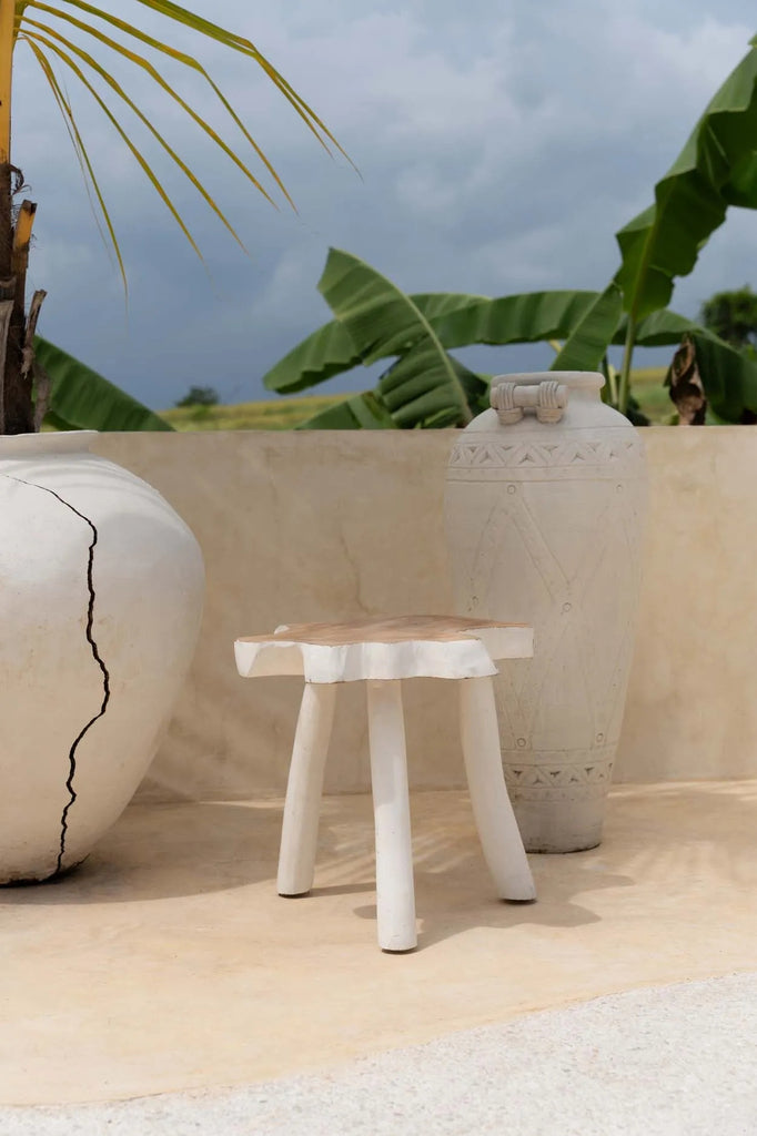 Witte keramische vaas en lage houten kruk op beige terras, omringd door groene bladeren en bewolkte hemel.