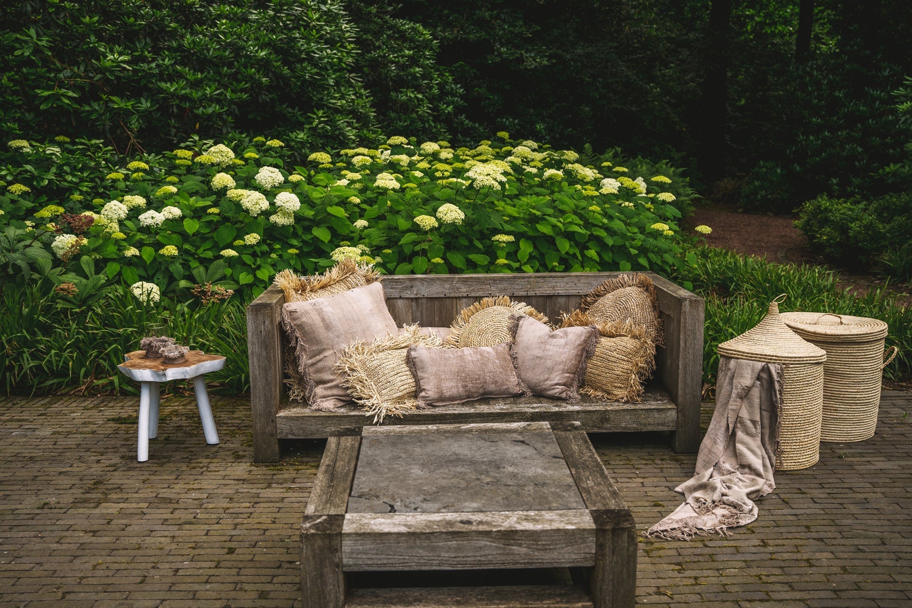 Houten tuinbank met beige kussens, naast een bijzettafel en rieten manden, omringd door groene planten.
