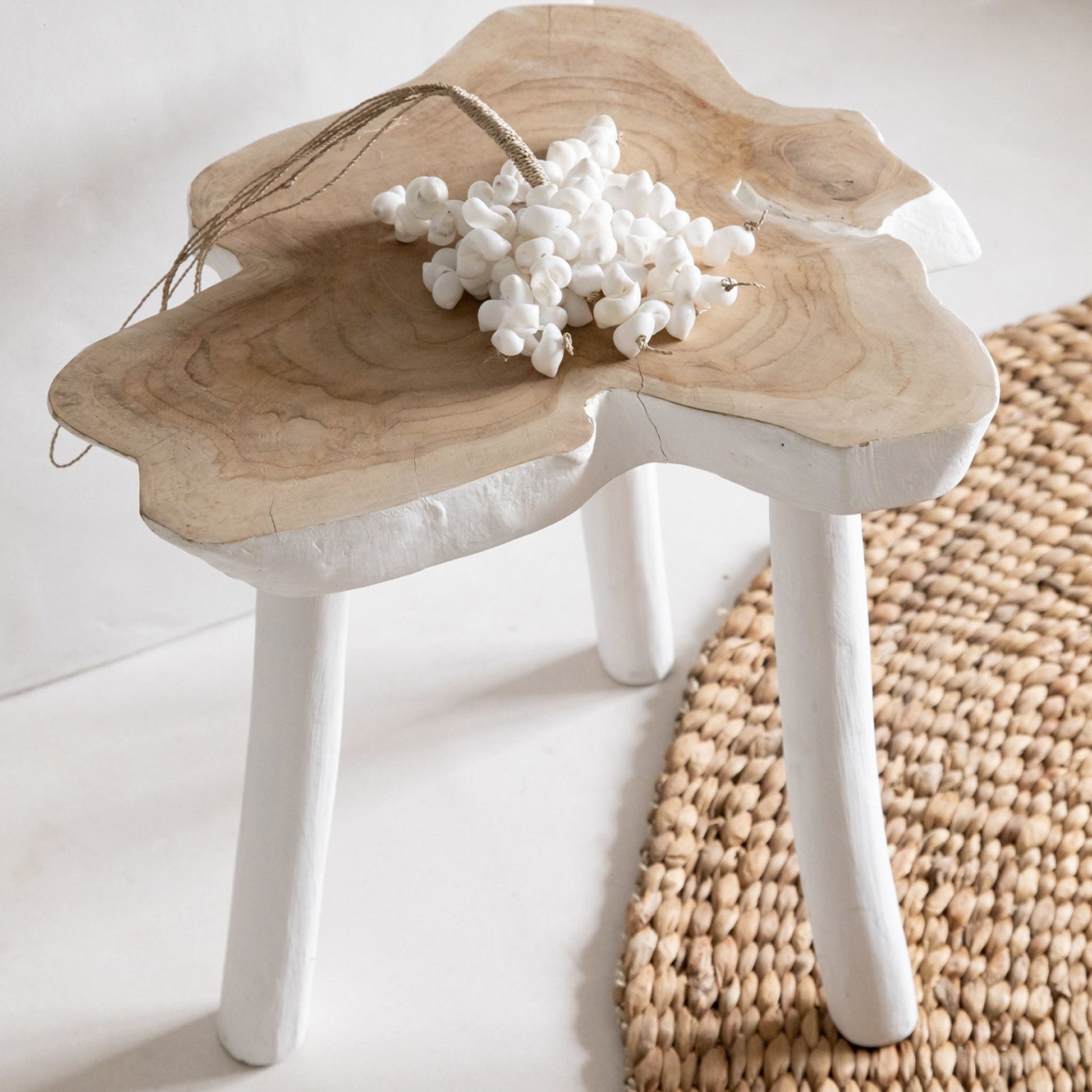 Houten kruk met natuurlijke vormen, versierd met witte bloemenkrans, op een geweven jute vloerkleed.