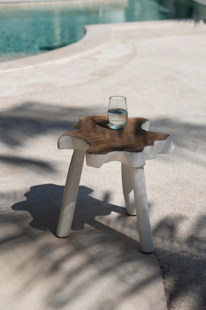 Glas water op een organische houten bijzettafel bij een zwembad, belicht door zachte zonnestralen.