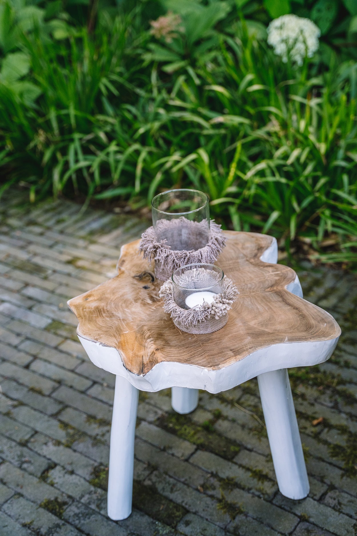 Rustieke boomstamtafel met witte poten, gedecoreerd met twee glazen windlichten met kaarsen in een tuin.