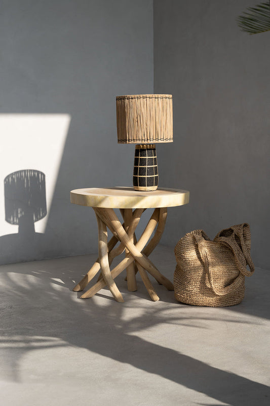 Houten lamp op gevlochten tafel, met rieten tas op grijze vloer in zonlicht, minimalistische setting.