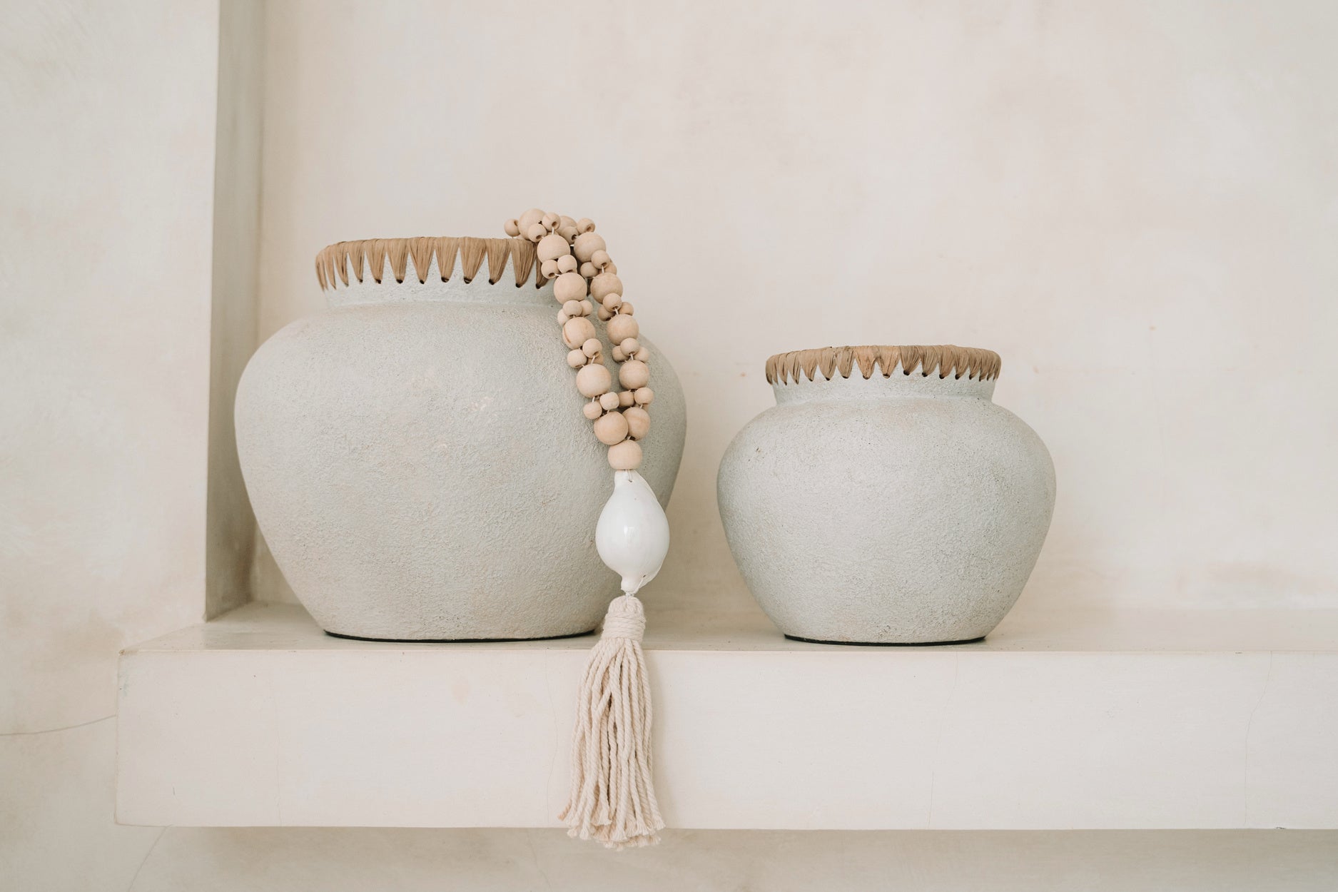Twee rustieke, grijze vazen met houten kralenketting op een witte plank tegen een beige muur.
