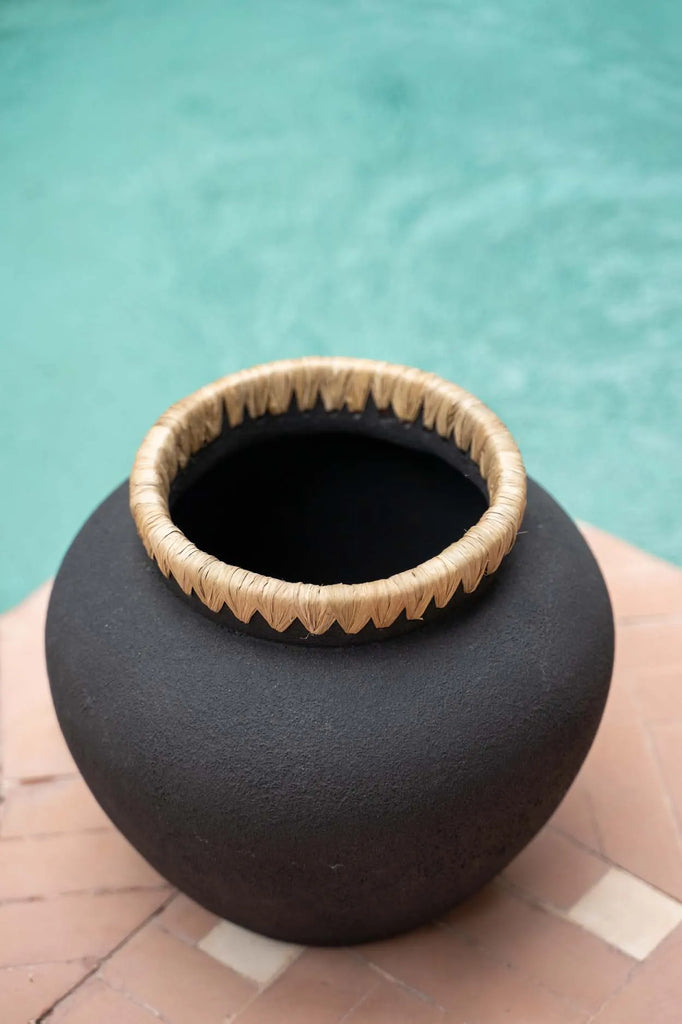 Donkerbruine kleipot met gevlochten rand, geplaatst op oranje tegelvloer naast helderblauw water.