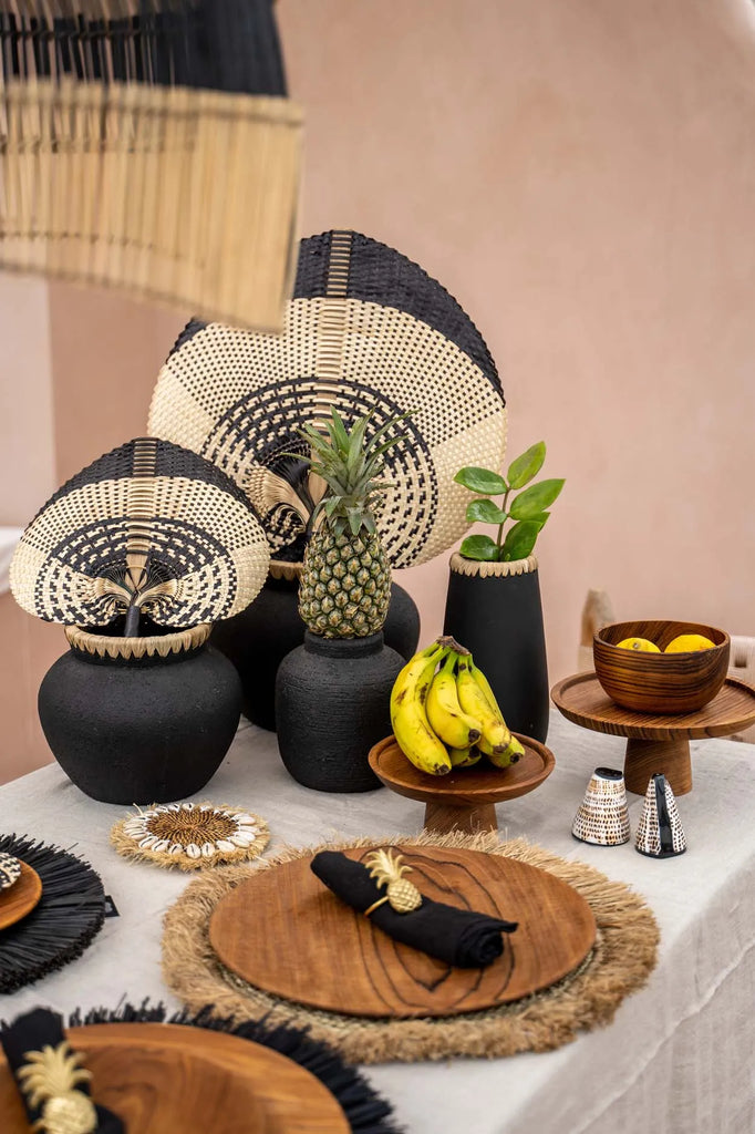 Rustieke tafelsetting met houten borden, zwarte vazen met vlechtwerk en decoratieve ananas en bananen.