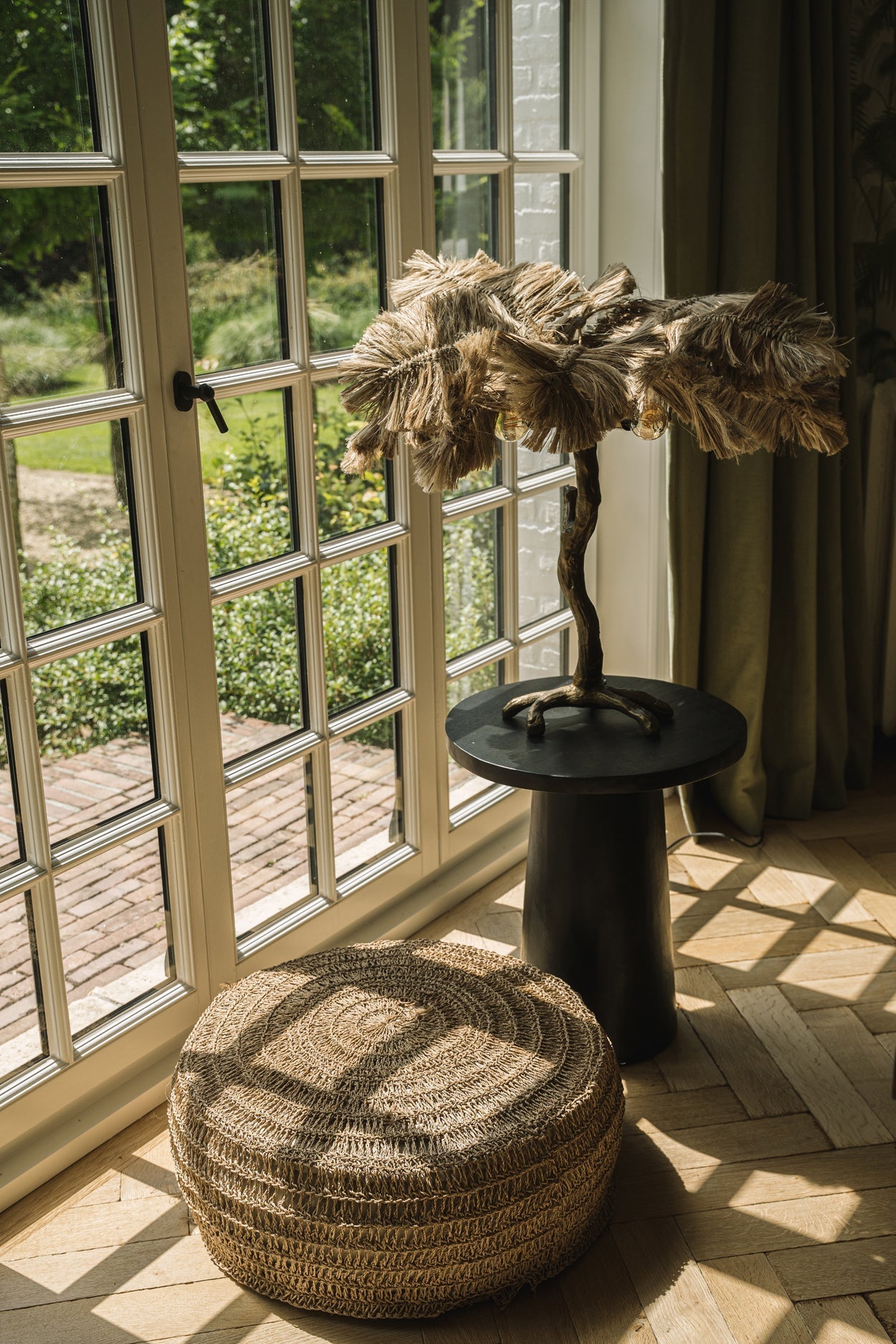 Rotan poef en rustieke lamp naast Franse deuren, zonlicht valt op houten vloer, groene tuin zichtbaar.