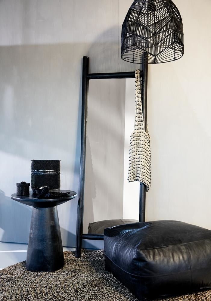 Minimalistisch interieur met zwarte leren poef, houten ladder met tas, bijzettafel en hanglamp op rustiek tapijt.