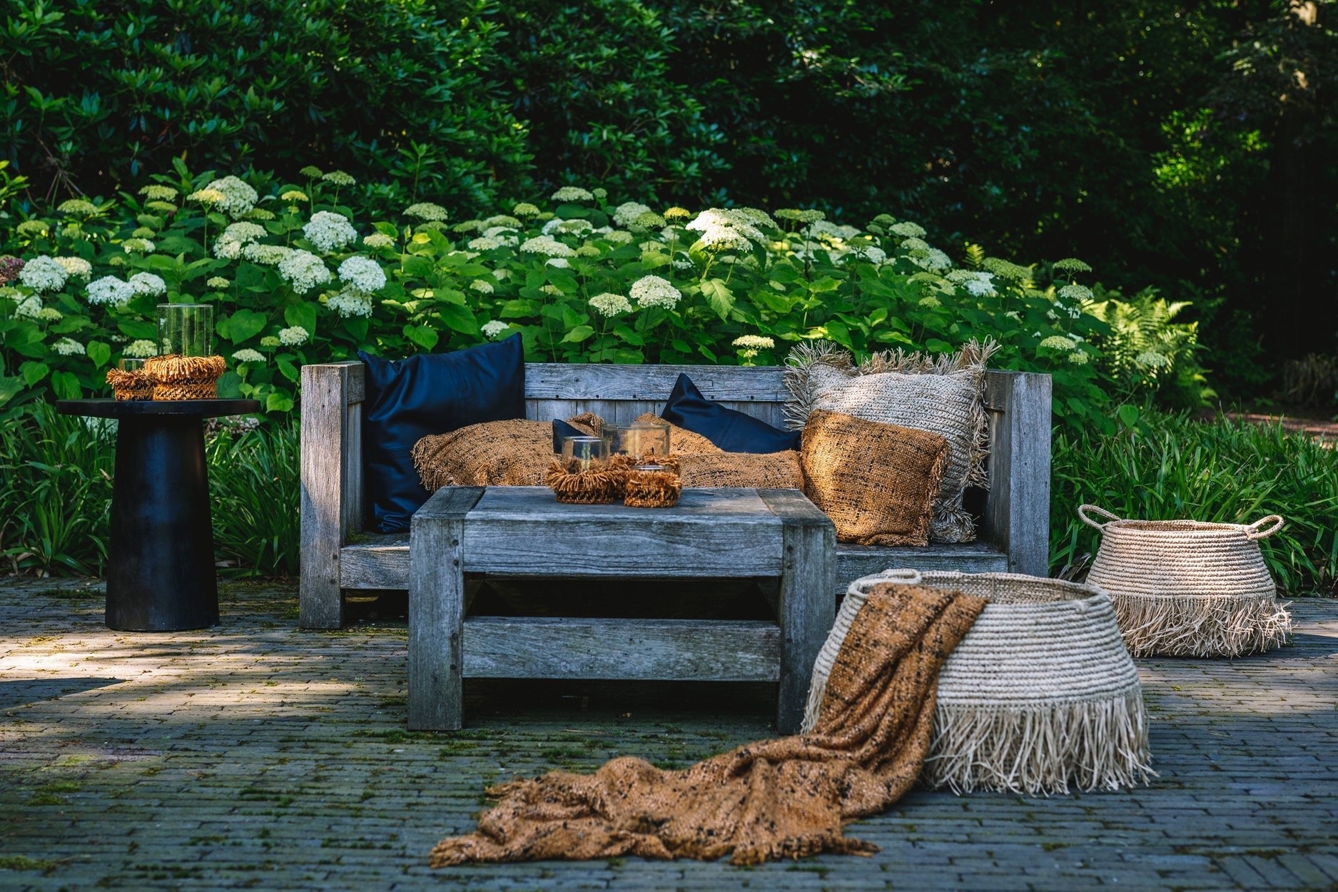 Rustieke houten terrasmeubelen met beige kussens en plaids, omringd door groene struiken en witte bloemen.