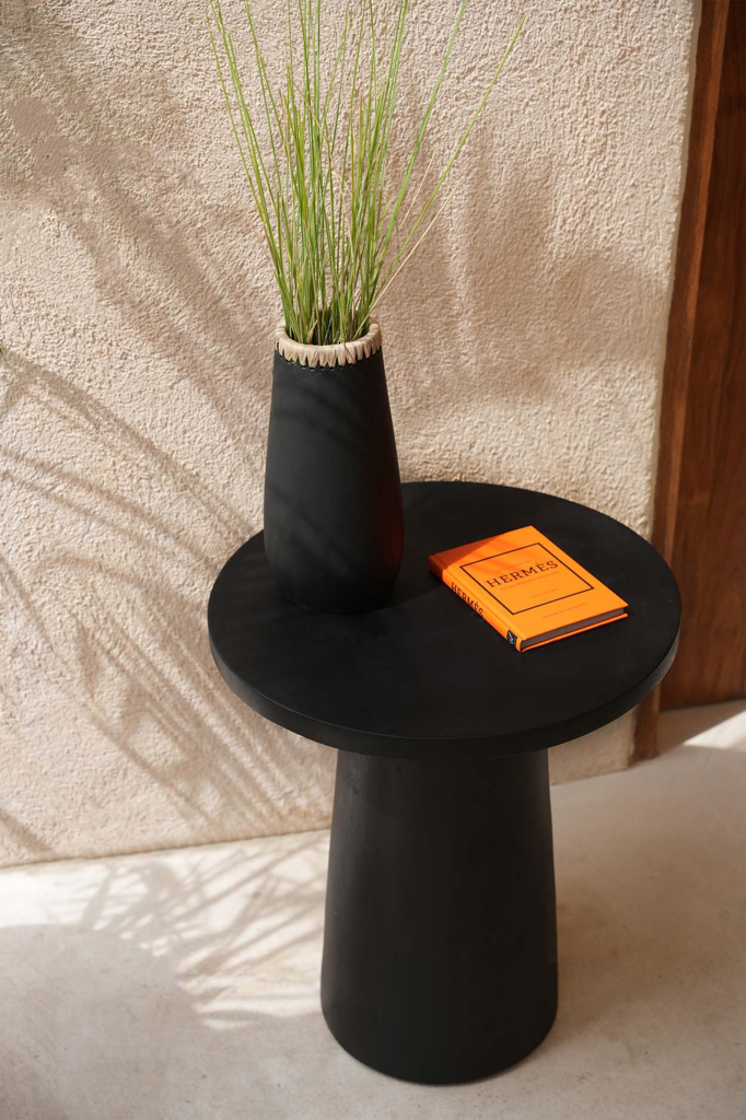 Zwart bijzettafeltje met hoge vaas groene grassen en oranje Hermès-boek, zonnige schaduw op muur.