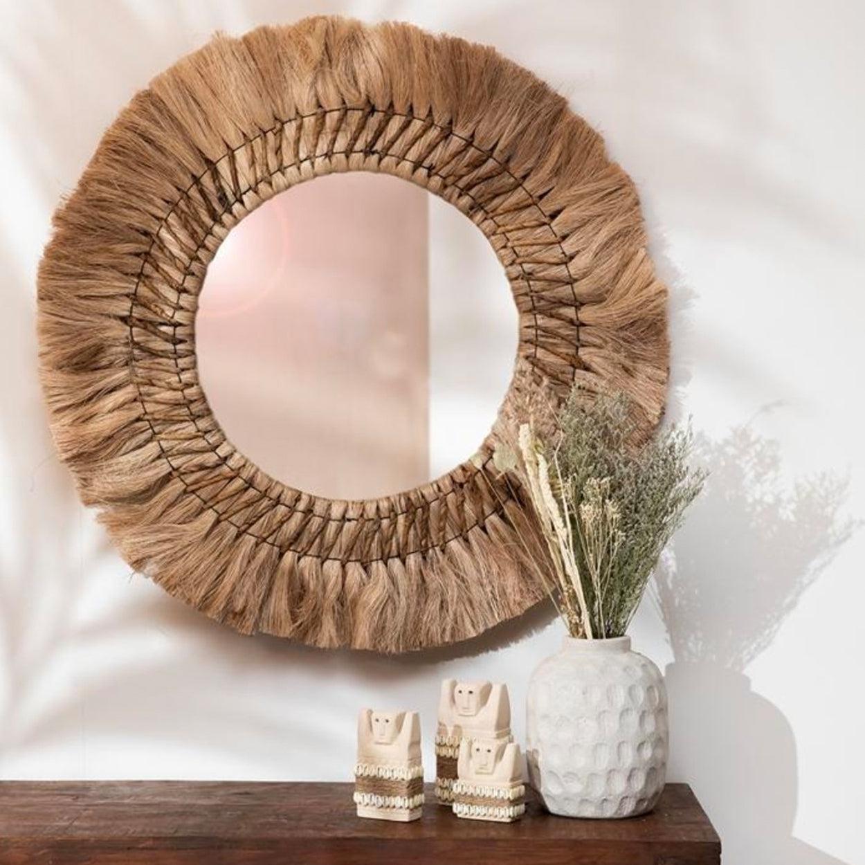Ronde spiegel met bruine raffia frame, witte vaas met gedroogde takken en drie kleine beeldjes op houten tafel.