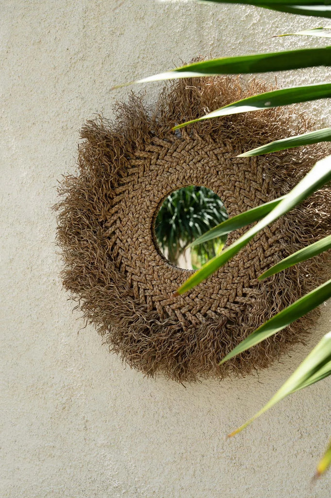 Ronde spiegel met gevlochten juterand op crèmekleurige muur, omlijst door groene bladeren.