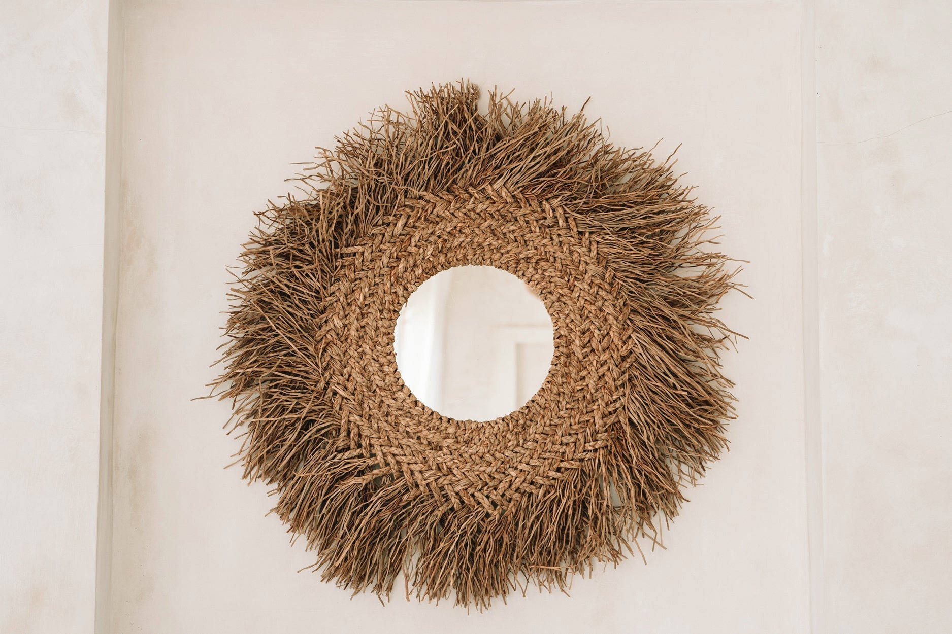 Ronde spiegel met een natuurlijke, gevlochten rotan omlijsting en ruwe textuur, tegen een lichte muur.