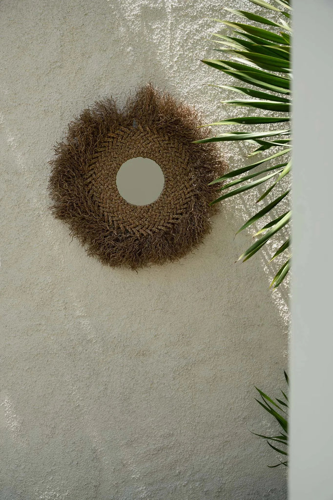 Ronde rieten spiegel hangt aan een beige muur, geflankeerd door groene palmbladeren in zonlicht.