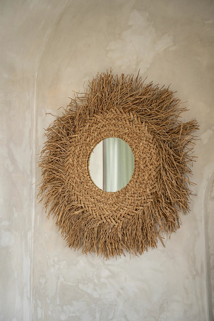 Rond spiegel met grof geweven rotan frame en spiraalvormige rand, hangt aan beige gepleisterde muur.