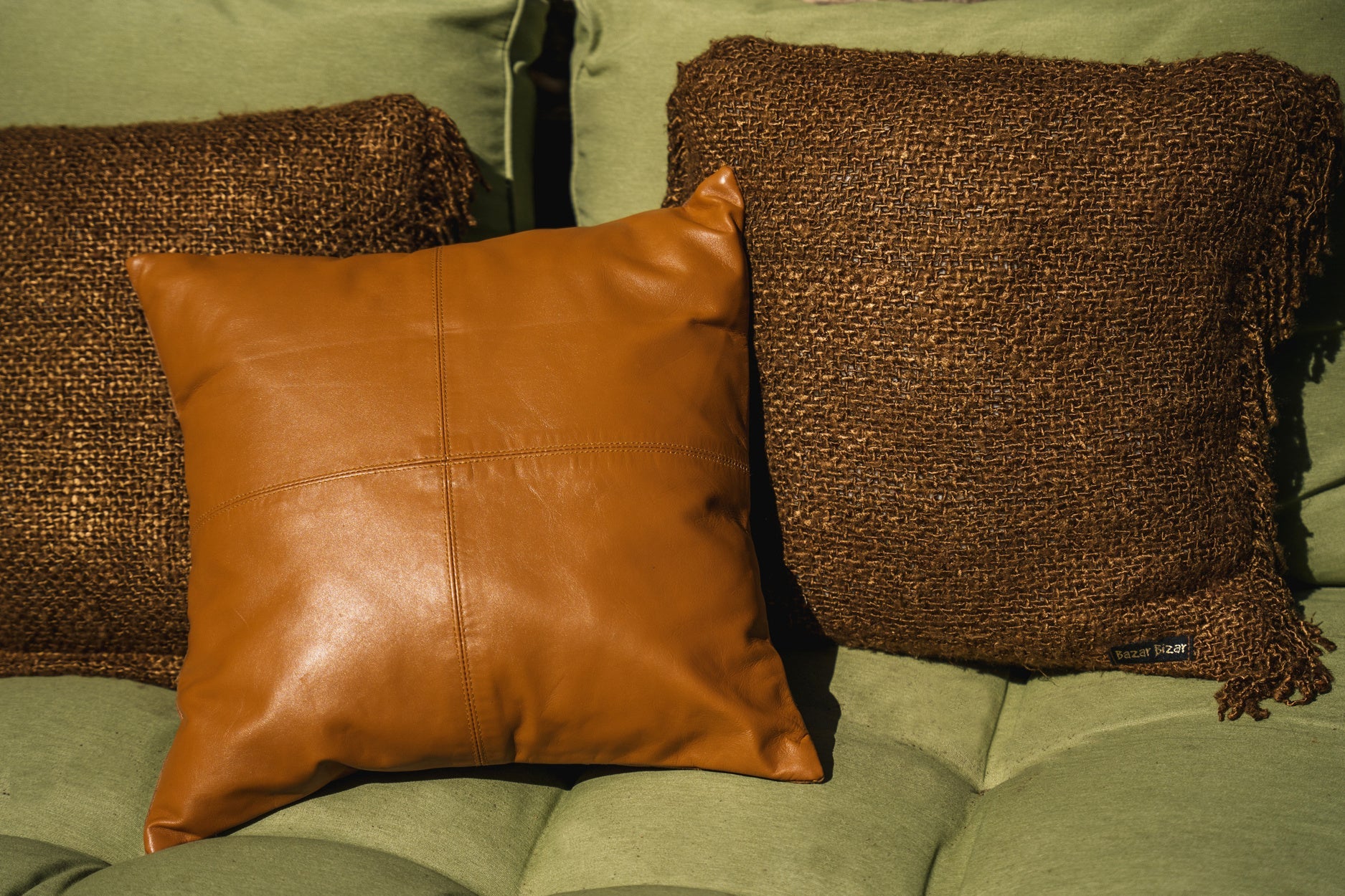 Bruin leren kussen naast bruin geweven kussen met "Bazar Bizar"-label op groene fauteuil.