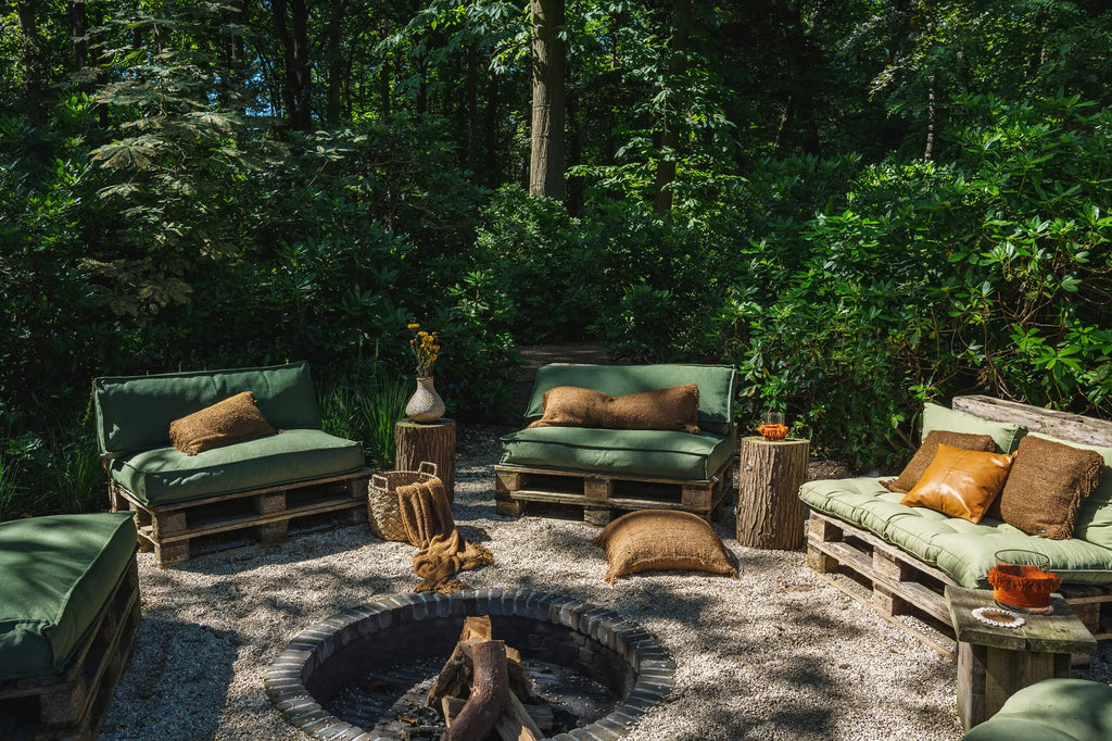 Groen ingerichte buitenlounge omringd door bomen, met houten meubelen, kussens en een ronde vuurplaats.
