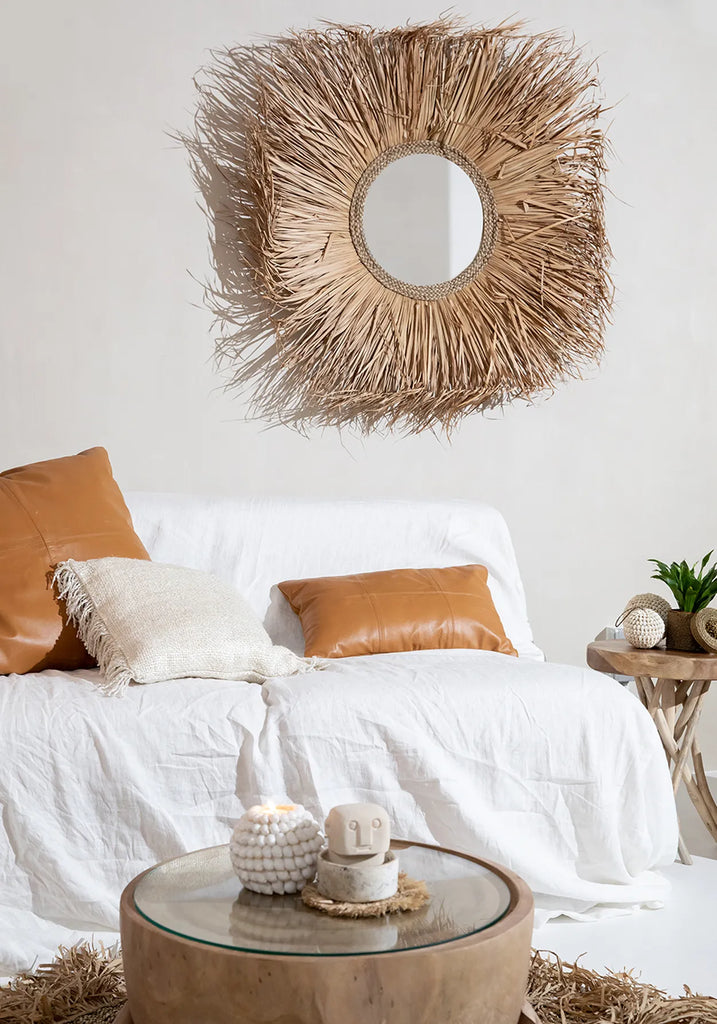 Modern woonkamer met rotan spiegel, witte bank met leren kussens, en houten salontafel met kaarsen.