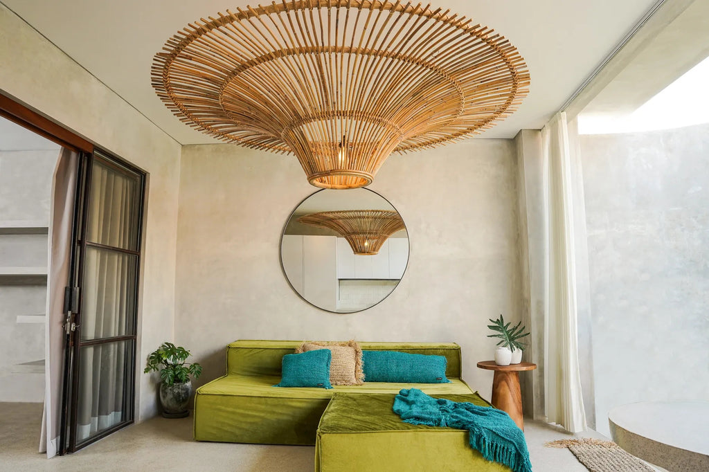 Stijlvolle woonkamer met grote rotan hanglamp, groene bank, turquoise kussens en ronde spiegel op lichte muur.