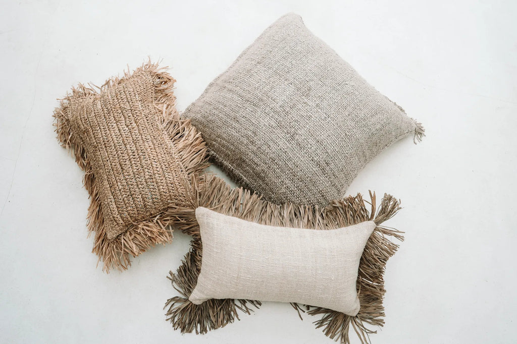 Drie decoratieve kussens met natuurlijke tinten en franjes op een lichte achtergrond, textuur en stijl benadrukt.