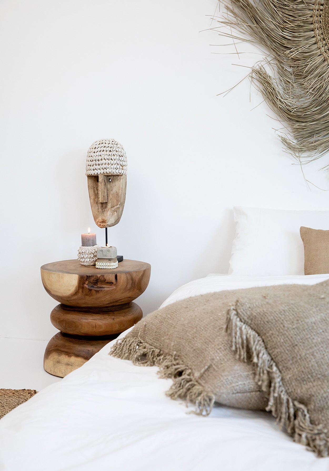 Rustieke slaapkamer met houten nachtkastje, schelpenmasker, kaarsen en naturel kussens op een wit bed.