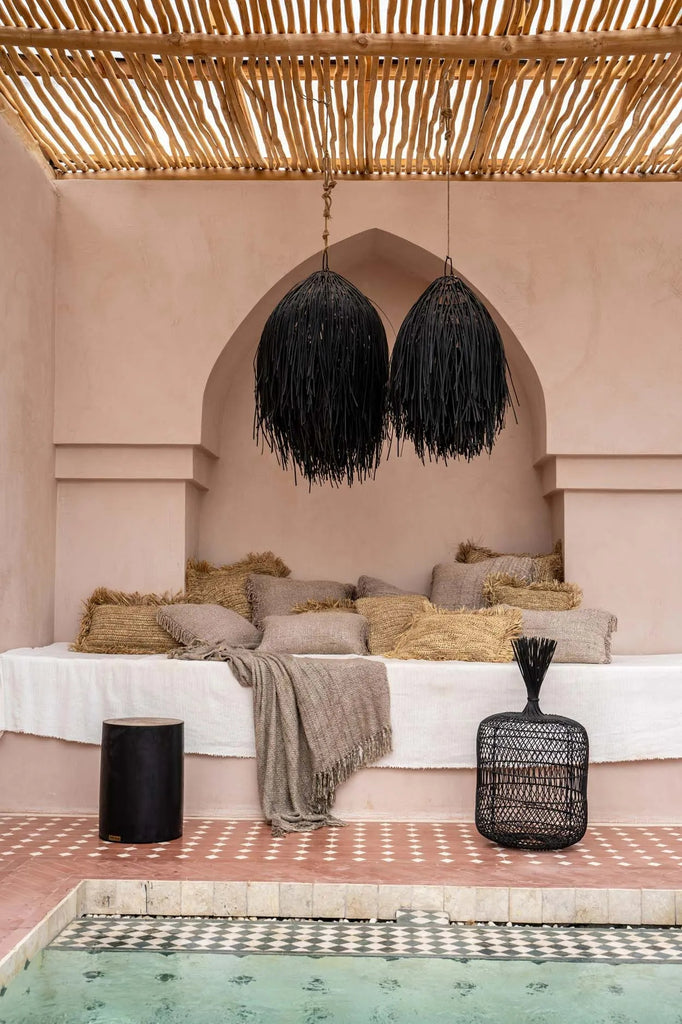 Knus loungehoekje met kussens en dekens, onder een houten lattenplafond met hanglampen bij een zwembad.