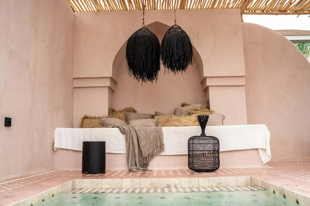 Rustieke loungehoek met kussens en zwart rotan hanglampen boven zonnig zwembad in beige resortomgeving.