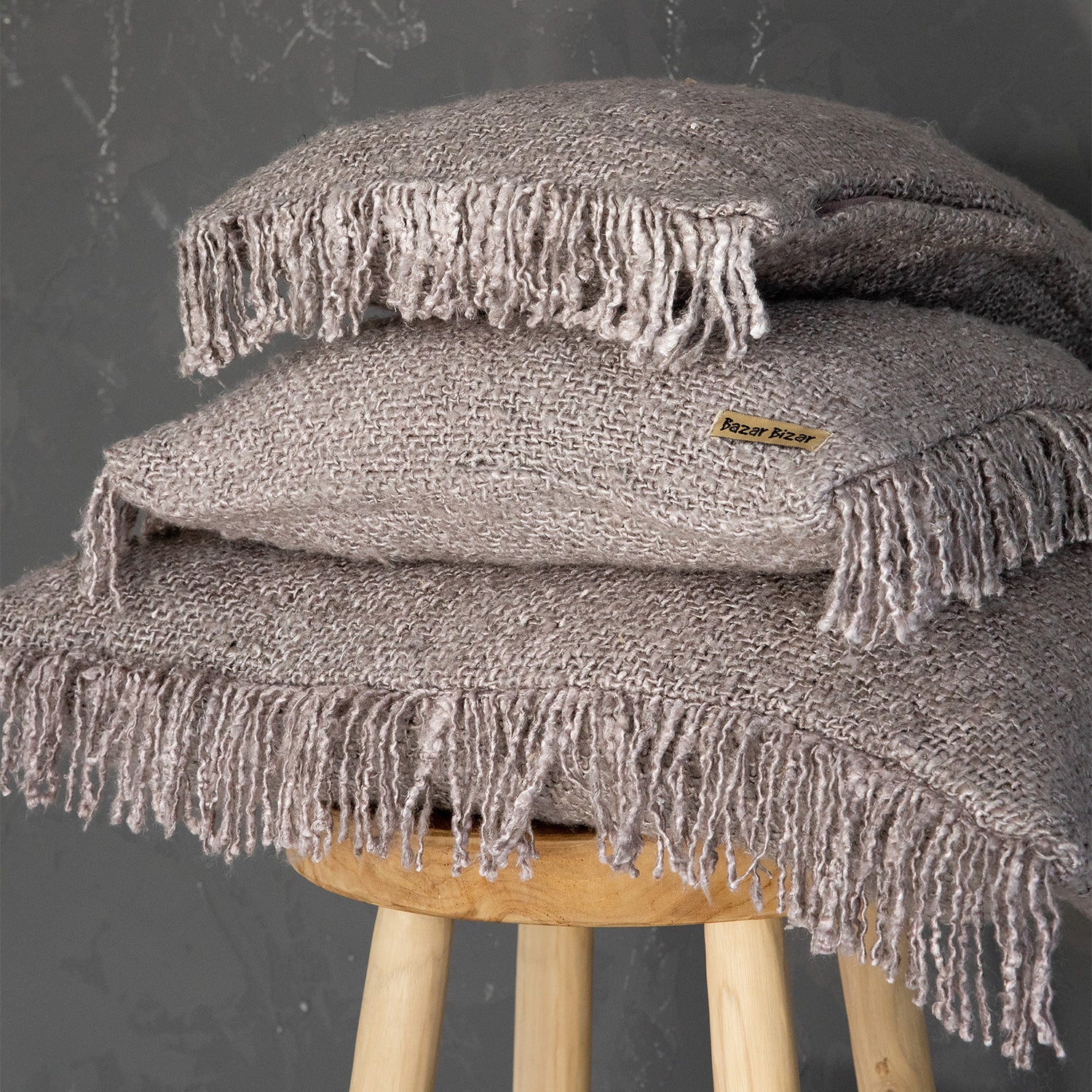 Twee beige kussens met franjes op houten kruk, label "Bazar Bizar", grijs betonnen muur op achtergrond.