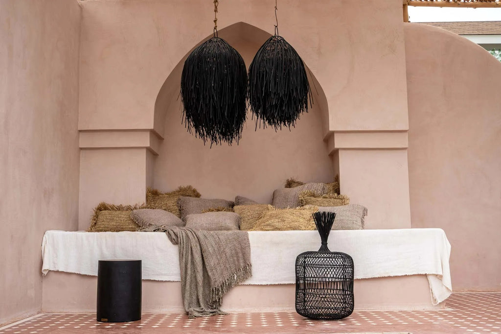 Gezellige hoek met lichtroze muren, beige kussens en een zwart gevlochten lamp boven de bank.