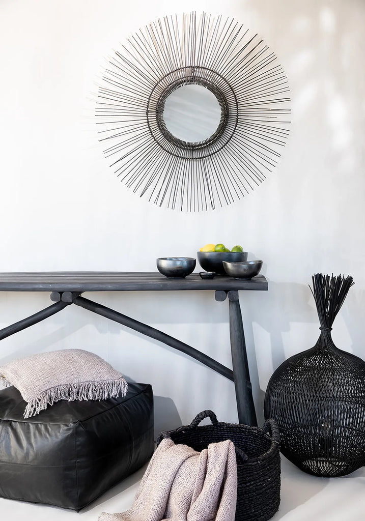 Wandspiegel met stralenontwerp boven console met fruitkommen, zwart poef met deken en mand in moderne kamer.