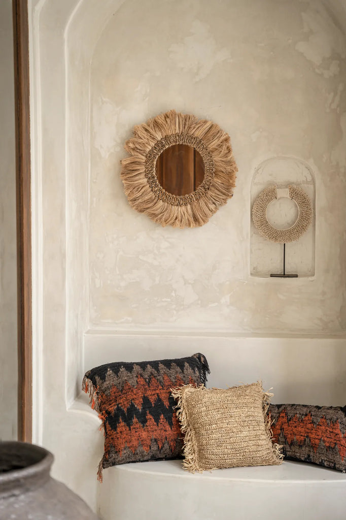 Rustieke muur met een spiegel met jute lijst, rieten mandala en boho kussens in warme aardetinten.