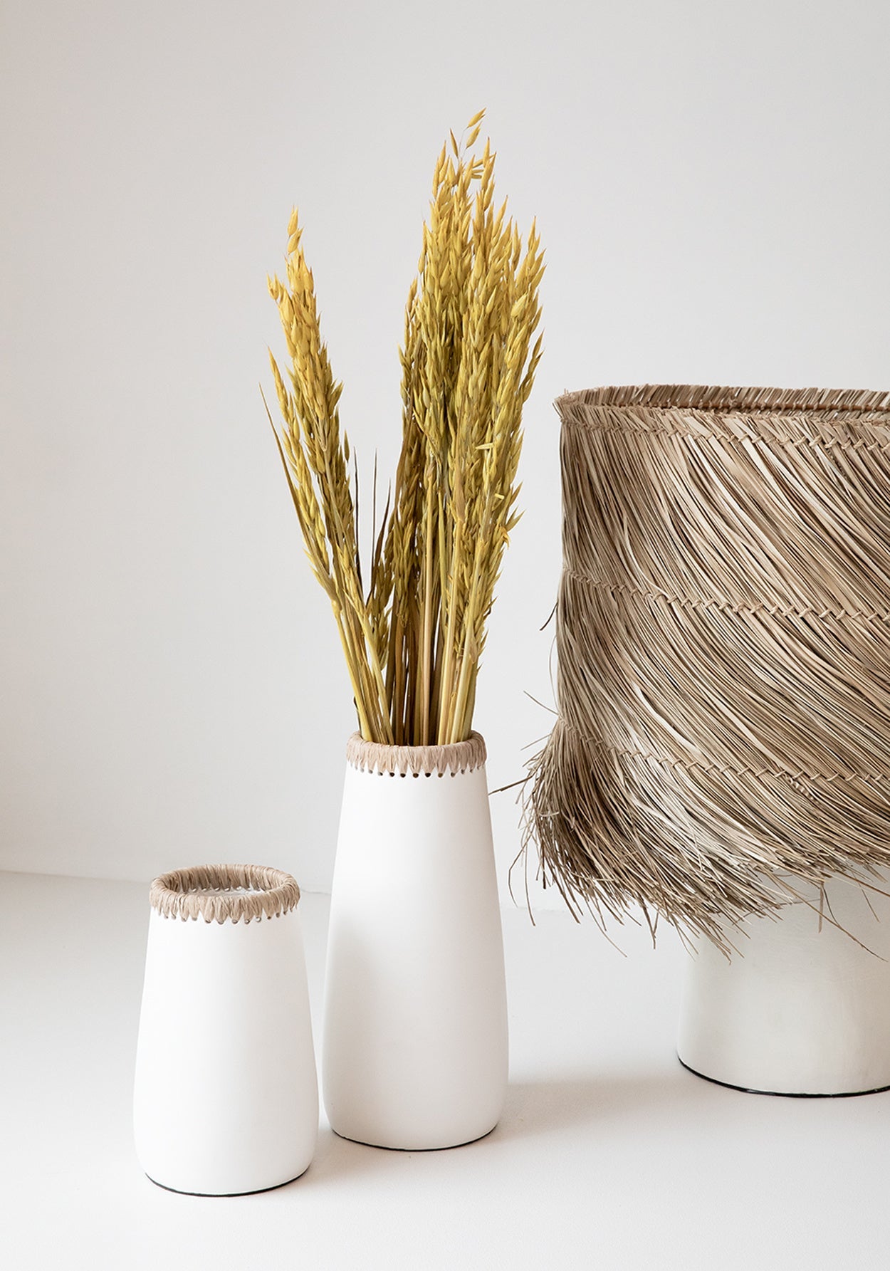 Witte vazen met gedroogde tarwearen en een mand van gevlochten raffia, op een minimalistische achtergrond.
